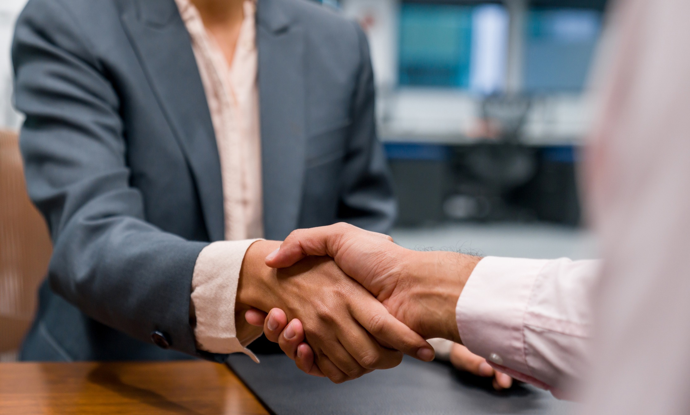 Two people in formal attire shaking hands over a desk in an office setting. One person is wearing a dark suit jacket, the other is in a light-colored shirt. The background is out of focus, emphasizing the handshake.