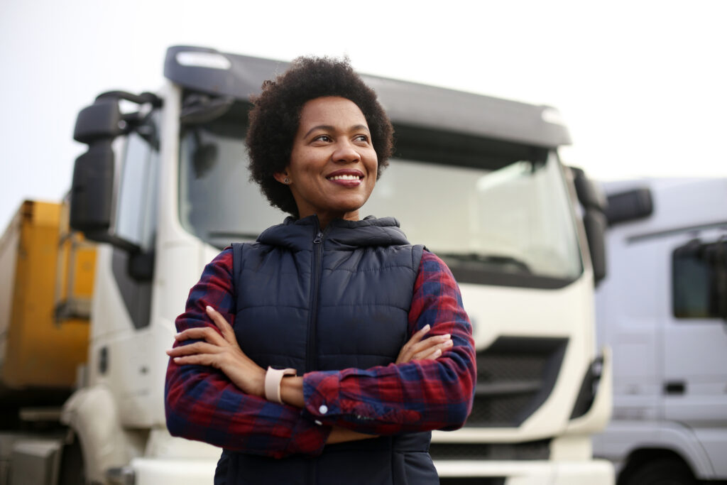 A person with short curly hair and a plaid shirt under a puffy vest stands confidently with crossed arms in front of a row of large trucks. The sky is overcast, adding a soft light to the scene.
