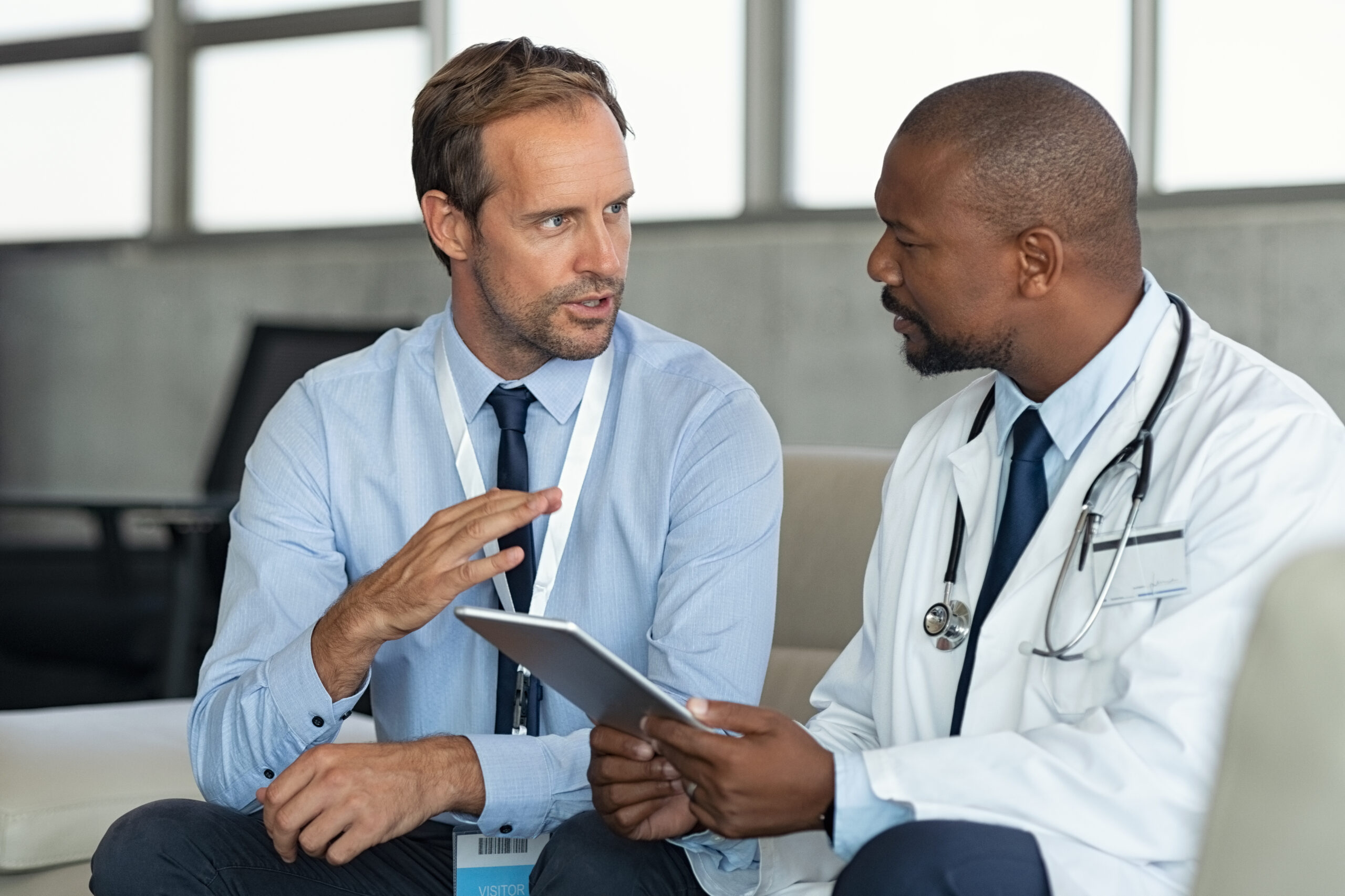 A doctor in a white coat with a stethoscope discusses something on a tablet with a man in business attire, who is gesturing with his hand. They are seated indoors and appear to be having a serious conversation.